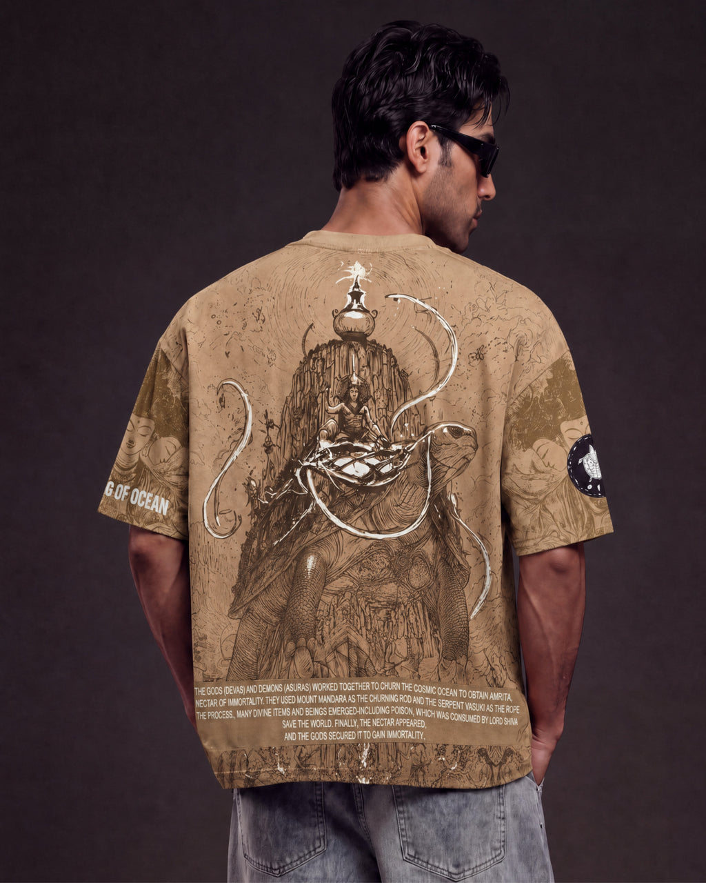 A man viewed from behind wearing a tan graphic t-shirt with a detailed illustration of a deity and Sansar, paired with light blue jeans against a dark studio background.