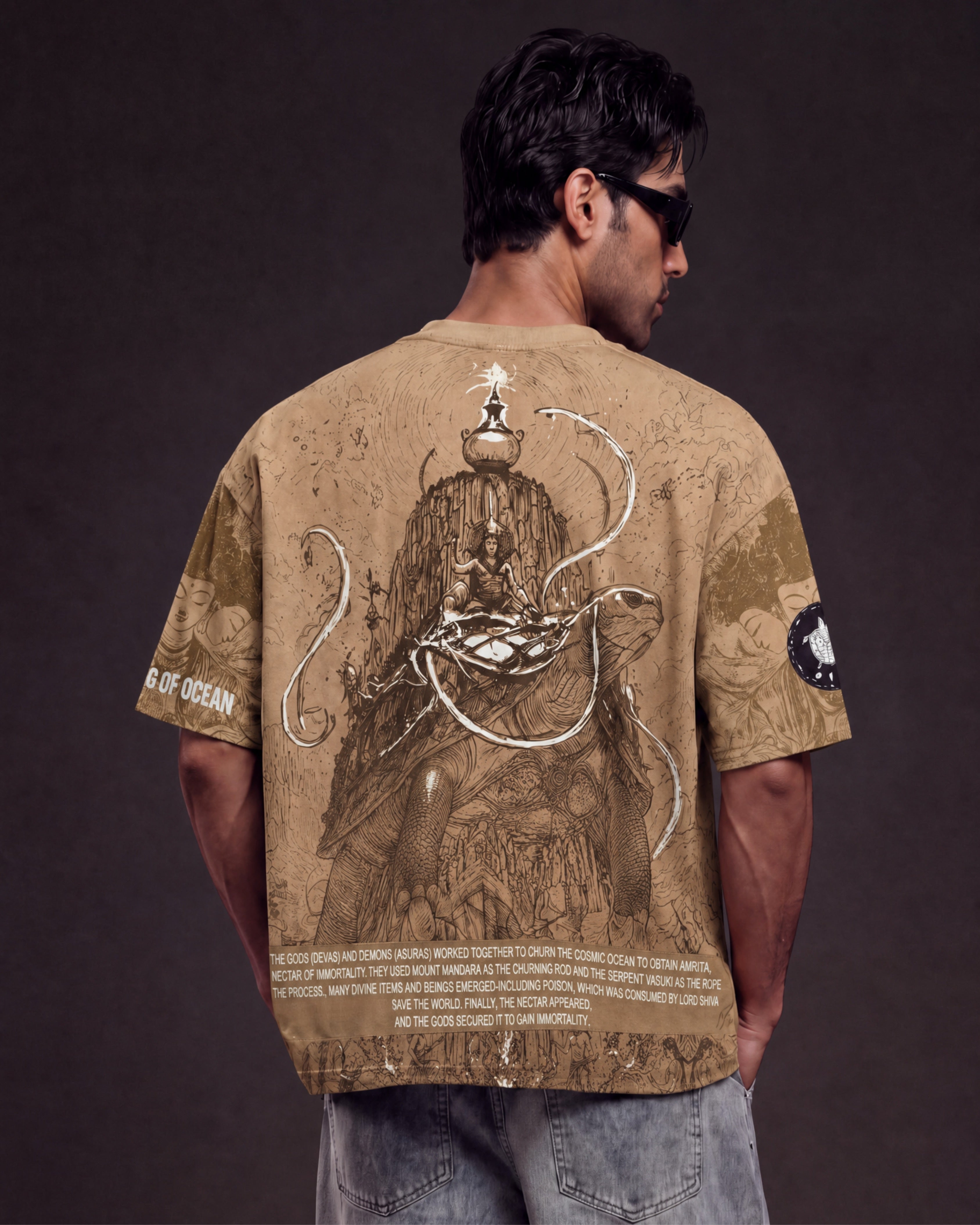 A man viewed from behind wearing a tan graphic t-shirt with a detailed illustration of a deity and Sansar, paired with light blue jeans against a dark studio background.